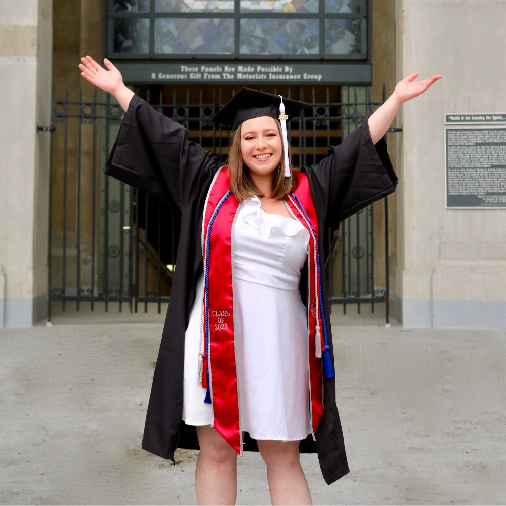 Image of young woman with her arms in the air wearing a black graduation cap and gown with a red stole and red, white and blue cords. Her tassel is white and says 2023. She smiling and wearing a white knee-length dress under her robe. Picture is in front of a stone building with black iron gates.