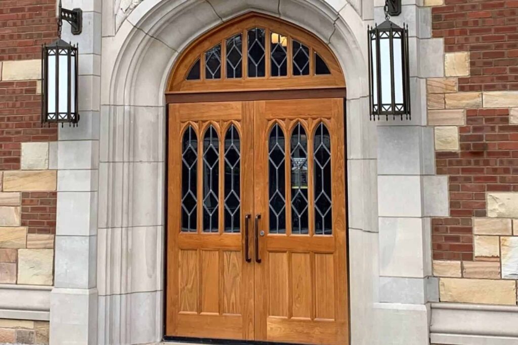 Close up of an arched doorway on a brick building. Double oak doors with glass windows with diamond patterns. A stone archway surrounds the doors and 2 large black lanterns are hanging on either side.