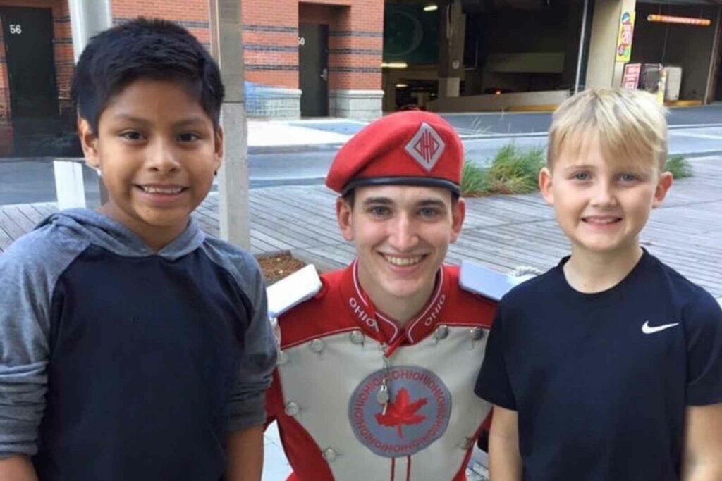 Two young boys on either size of a college drum major. Boys are standing on a sidewalk in front of a brick building and parking garage. Young man is crouched down between them. He is wearing his bank uniform