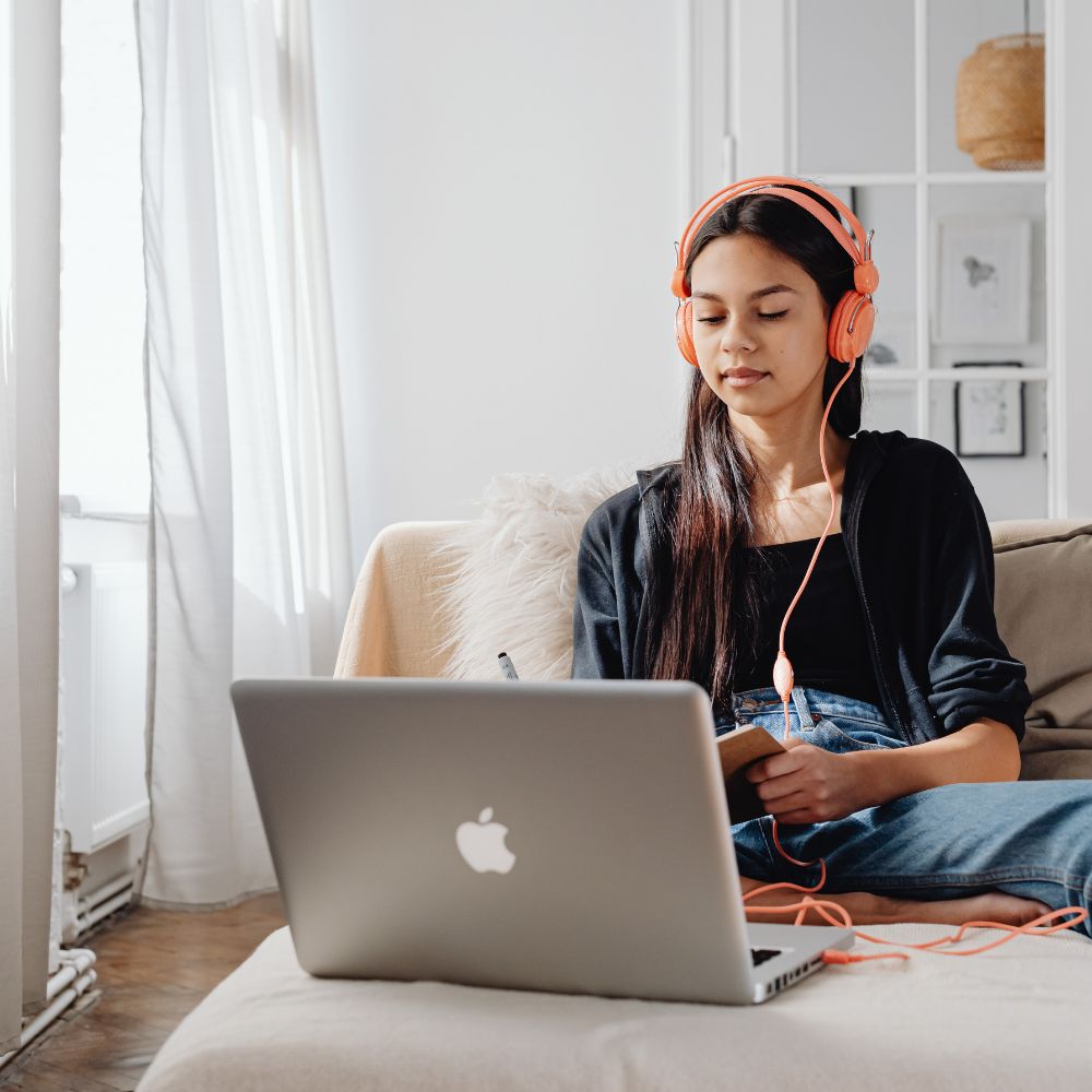 College student sitting in a room with white walls, a french door and white curtains. She's sitting on a cream colored counch and her Macbook is open on a large cushion. Girl is facing the camera but looking down at what she's writing on a notepad. She's wearing a black hoodie and jeans with tangerine colored headphones and cord.