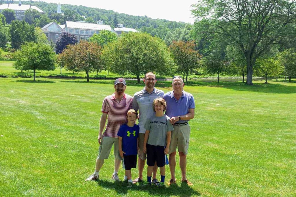 Three men and 2 boys standing in a large green space, facing and smiling at the camera. Behind them are trees surrounding a pond and behind that are large campus buildings.