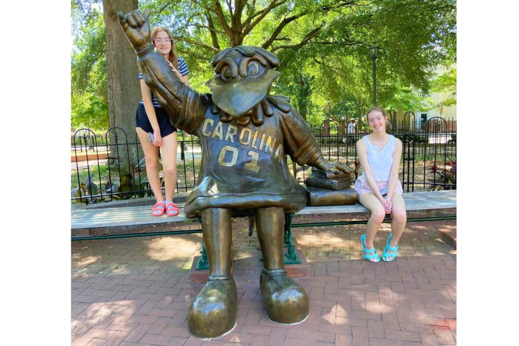 Two girls stand on either side of a statue of the University of South Carolina mascot. One is on the left, standing on a bench and leaning on the statue's outstretched arm. The other girl is on the right, sitting on the bench next to a stack of books.