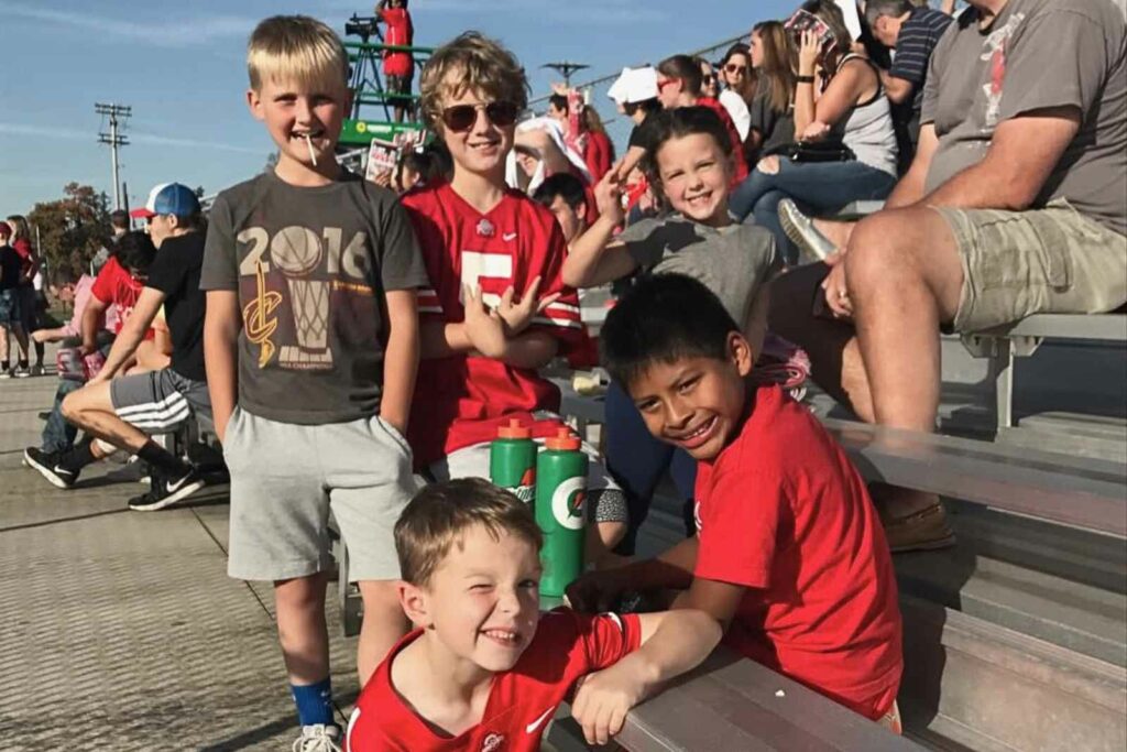 5 kids on bleachers in the bright sun. Bleachers are full of a lot of people. Four boys and 1 girl are facing the camera and smiling.