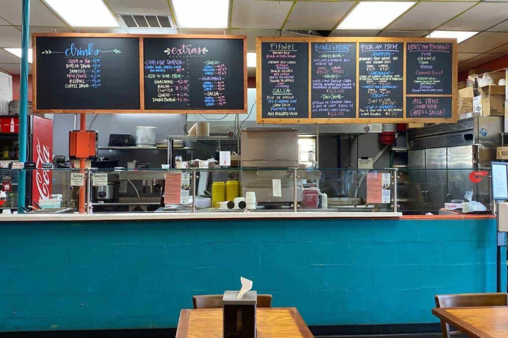 Interior of a burrito restaurant facing the counter. Wall beneath the counter is painted turquoise and the counter is protected by a glass shield. A Coca Cola machine is on the left and a silver industrial cooler is on the right. Hanging from the ceiling are chalkboards with the menu handlettered in various colors of chalk.