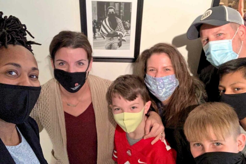 7 people wearing masks and facing the camera. Posing in front of a black and white photo of a man with an American flag draped across his shoulders.