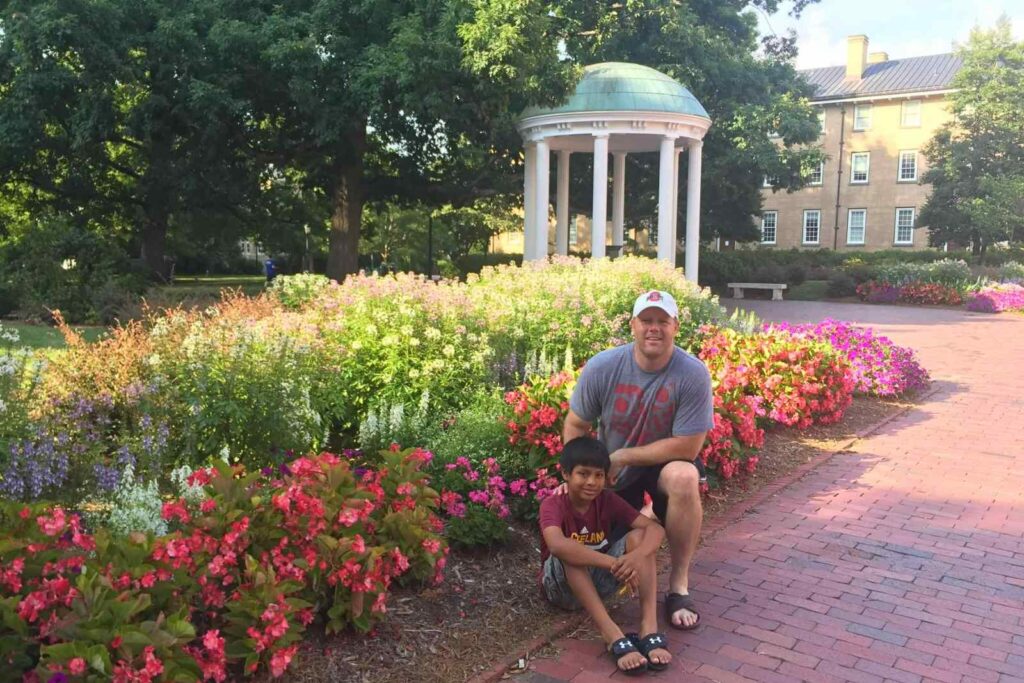 A man wearing a white baseball cap and gray tshirt is kneeling next to a boy in a burgundy tshirt who is sitting next to a red brick path. Behind them are flower gardens with shades of pink, purple and red. Behind the flowers is the Old Well at UNC.