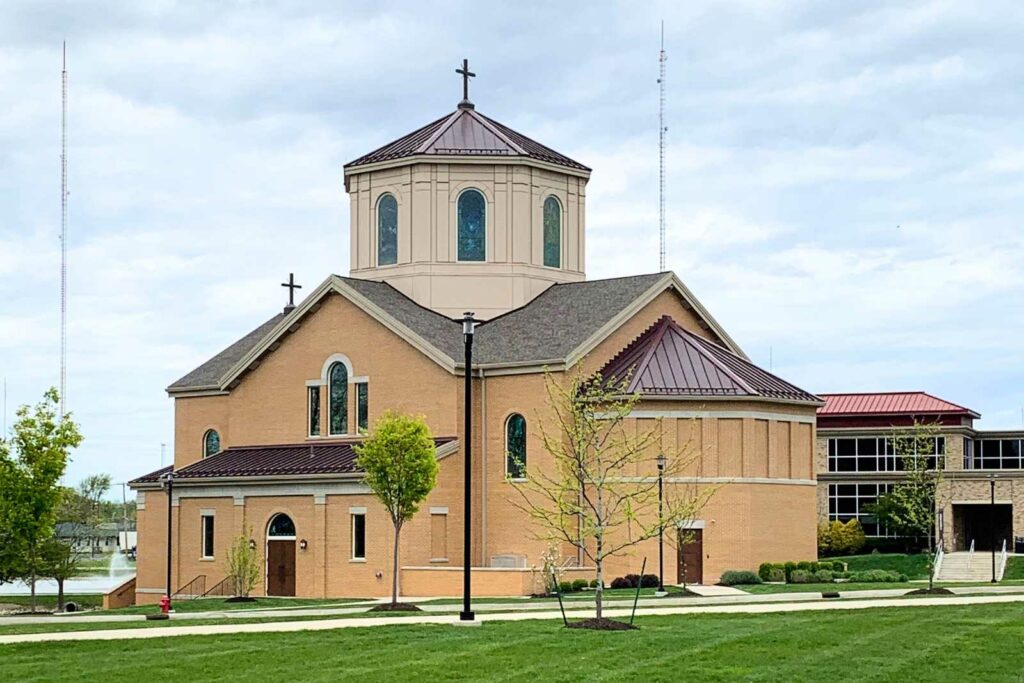 College chapel with green grass in foreground and a few newly planted trees with a blue cloudy sky behind. Building is tan brick with stamped metal burgundy rooftops and 2 crosses at the top of the chapel.