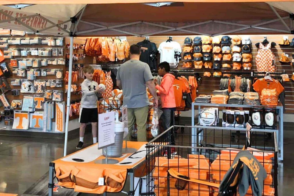 Interior shot of a college bookstore. A man and 2 boys are looking at basketballs in a bin, surrounded by racks of hats, tshirts, car decals in bright orange, black and white.