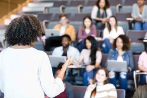 A close up of the back of a black female professor wearing a white blouse and gesturing to her students. Students are blurred in the background, facing her and sitting in a lecture hall with laptops or notebooks on their desks.