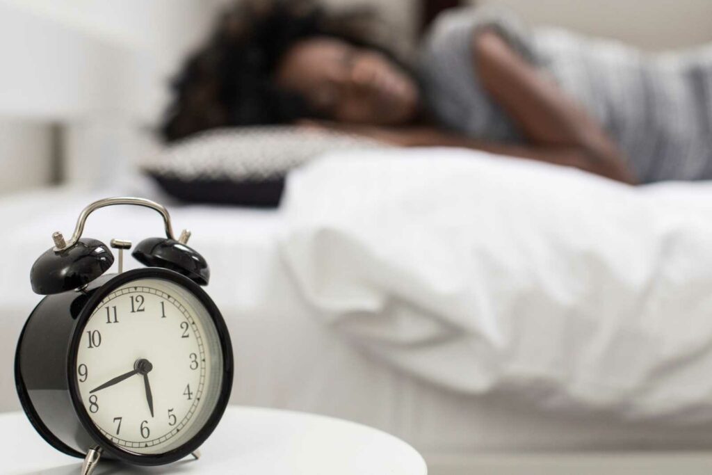 Blurred background is a Black woman wearing a gray tshirt laying on a pillow on a bed with white sheets and a white duvet. A nightstand is on the left side in the foreground. Resting on the white nightstand is a vintage black alarm clock