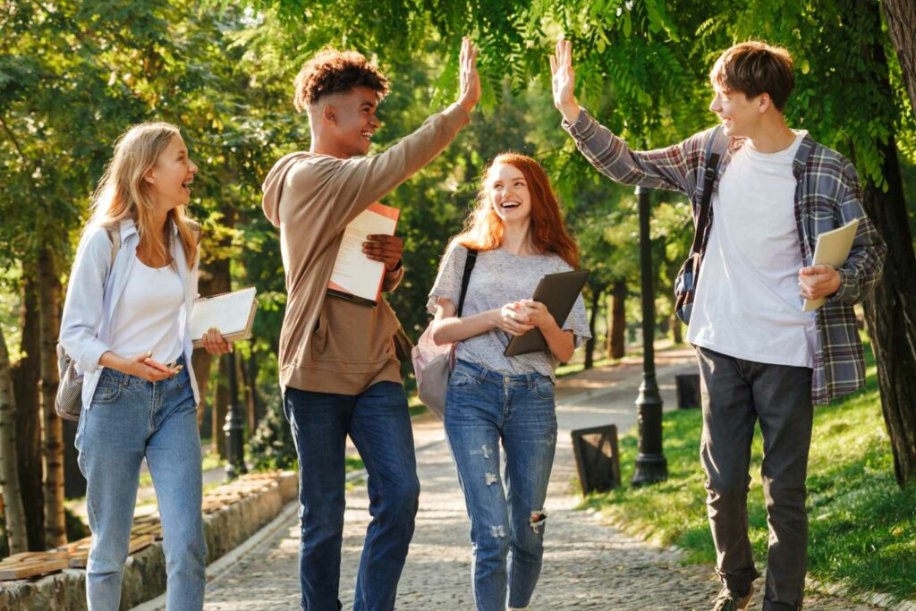 Four college students walking on a path with green grass, trees and light posts on both sides. Two young men wearing jeans are in the front walking toward the camera but facing each other while doing a high five. Two young woman are laughing and walking behind them.