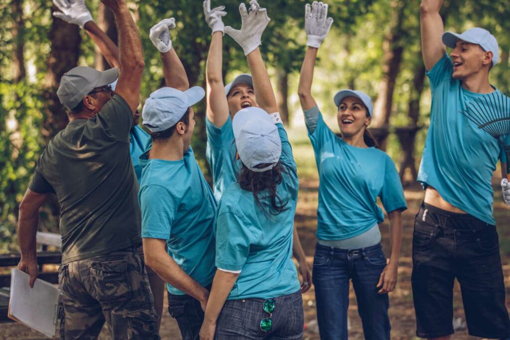 Group of 7 young adults in a clearing in the woods, raising one arm in the air as if they just broke a huddle. 6 are wearing plain teal tshirts and light blue ball caps. One man is wearing an olive tshirt, tan cap and camo pants.
