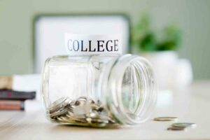 A jar is turned on its side and partially filled with silver coins. A few coins are on the white table. A white piece of paper is on top of the jar that says "COLLEGE" in black letters. A blurred background is made up of books, a laptop and a plant.