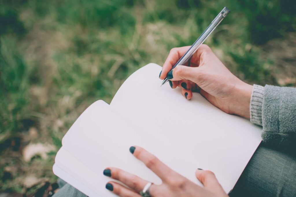 Close up of a blank journal with plain white pages is open on a woman's lap. Woman is about to write on the page with a silver pen. She has dark painted nails and is wearing jeans and a long sleeve gray sweater. Grass is blurred in the background.