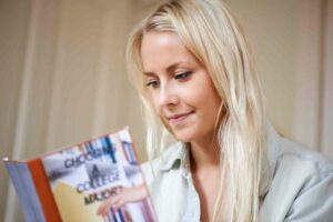 Young woman with long blond hair and a faded denim collared shirt is looking at a brochure with the title "Choosing a College Major"