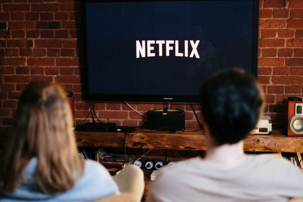 Large flat screen tv on a brick wall above a wooden console. Screen is black with Netflix in large white letters in the middle. The back of a woman and a man facing the tv are blurred in the foreground.