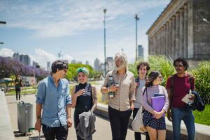 A group of 6 college students walking toward the camera. Students are outside with campus building behind them and a city skyline in the background. They are carrying books, water bottles, jackets, and wearing backpacks. Students are a variety of ages, genders and races.