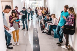 A group of college students standing in a wide well-lit hallway with white walls, a white floor with a black strip in the middle and a turquoise bench. Some students are sitting, others are leaning against the wall or huddled in a small group. They're looking on their phones, talking or sharing information on paper. Many are wearing backpacks or carrying bags.