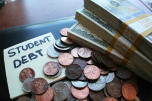 Close up photo of three stacks of 100 dollar bills wrapped in labels that say $10,000. A pile of coins is next to the bills and a few coins cover a post-it note pad with "Student Debt" written in black marker.