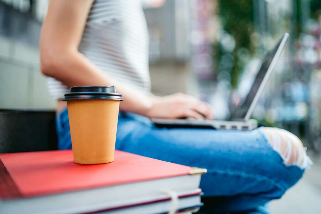 College student sitting outside on a bench wearing jeans ripped at the knee and a sleeveless top. Legs are tucked under her and she's typing on a laptop on her lap. She's out of focus in the background. In the foreground is a stack of books with a red one on top and then a to-go cup of coffee
