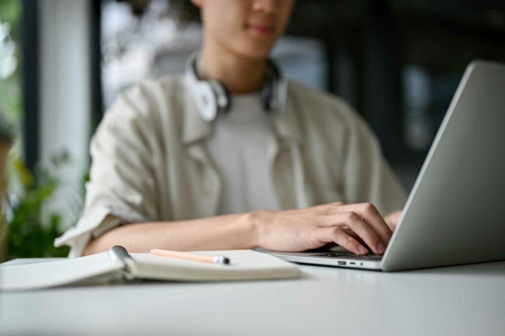 College student typing on a laptop on a desk. Student is facing the camera and out of focus. He's wearing a tan shirt with a white tshirt underneath. His silver headphones are around his neck. An open notebook with a pencil is on the table next to him.