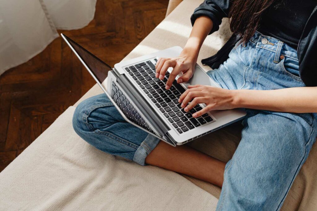 College student sitting on her bed and cropped so only her legs and part of her arms are visible. She has one leg folded toward the other leg with her laptop resting on top. She's wearing jeans and a dark navy top as she types. The blanket on her bed is beige.