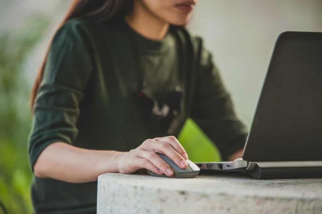 A college student wearing a dark green tshirt is facing a laptop and moving a mouse. The laptop is sitting on a concrete table. The college student is out of focus in the bakcground.