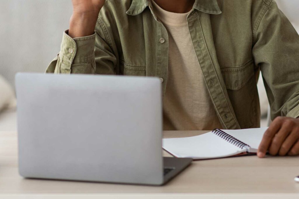 A college student is leaning on a light wooden desk, wearing an olive buttoned shirt with a beige tshirt underneath. His face isn't visible. He has a blank notebook open and is resting his hand on the pages.
