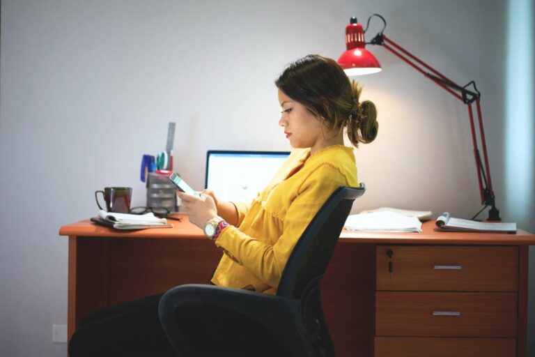 College girl sitting at her desk in her dorm room. Her desk hs facing a blank white wall and is covered with books, papers, a mug, laptop and a red lamp. She is turned in her chair and facing the left, looking at her phone. She's wearing a yellow top, sitting in a black desk chair and she has her hair pulled back.