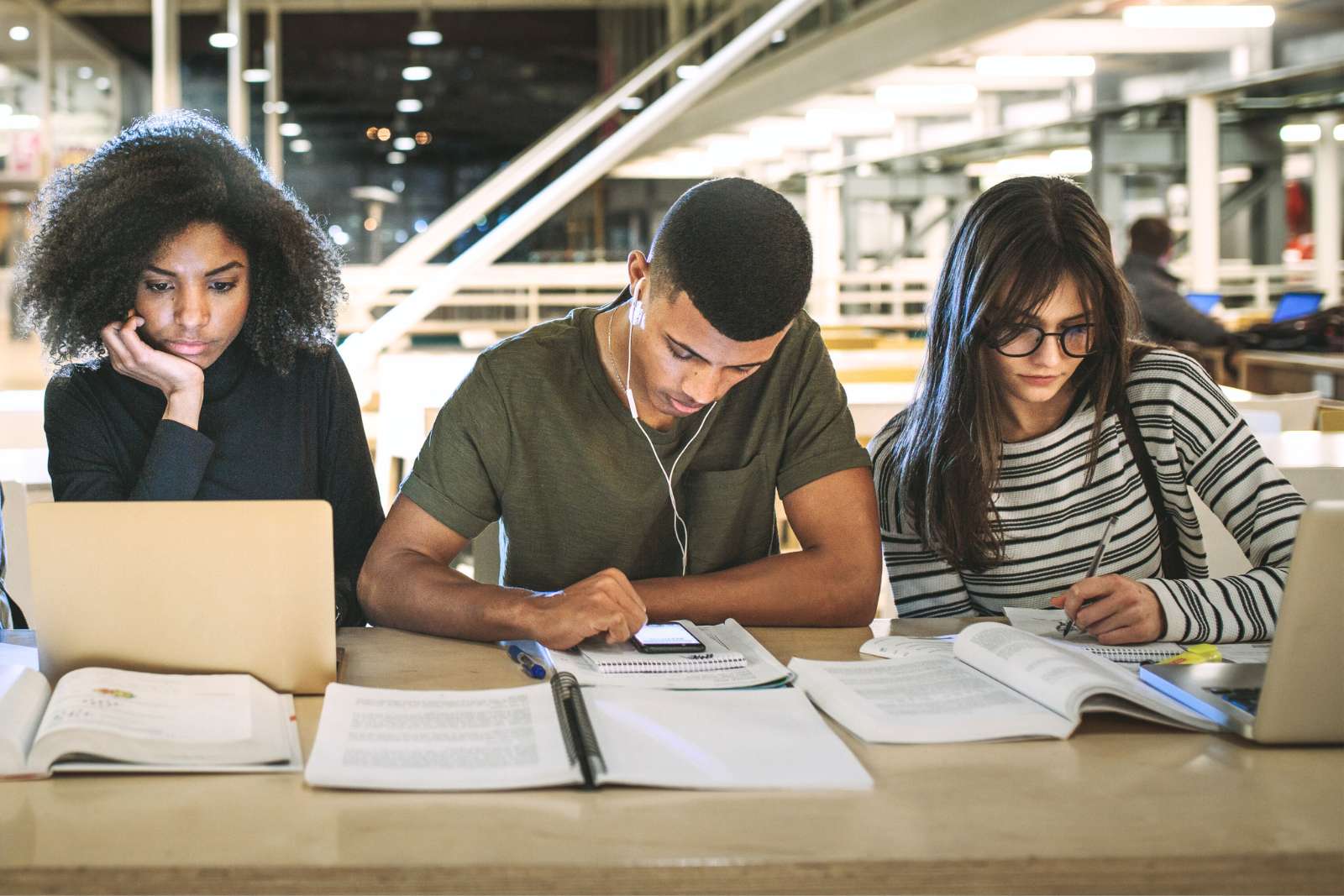 3 college students in a library sitting at a table facing the camera. They're studying. One student is looking at her laptop screen, one student is wearing earbuds and scrolling on his phone, the third student is writing in a notebook while looking at an open textbook next to her laptop.