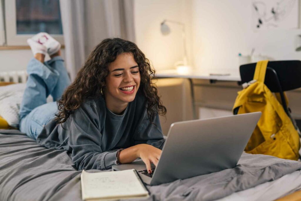 Female college students laying on her stomach on her bed. Her feet are kicked up behind her and she's propped on her elbows, using a laptop computer. She has long dark curly hair and is wearing jeans and a distressed gray top. A notebook is on the bed next to her laptop. A desk, lamp, black chair with yellow backpack over it are visible in the background.