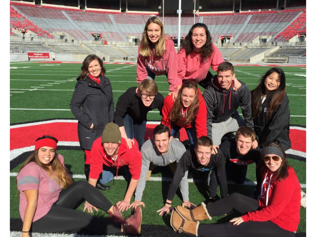 A group of college students and a faculty member making a pyramid on a football field. Everyone is wearing red, black and gray clothing.