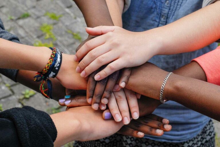 Image of teenager's hands reaching into the middle of a group and stacked on top of each other. A diverse group with different nail colors, bracelets, and clothing.